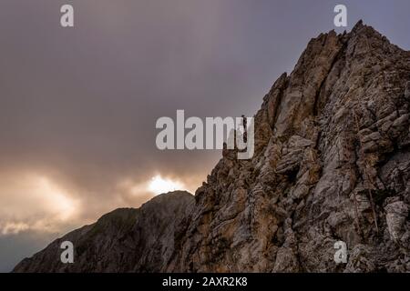 La jeune femme se tient sur un affleurement rocheux à Karwendel Ouest sur la route haute de Mittenwalder. Fond nuageux avec soleil en soirée et croix au sommet de Linde Banque D'Images