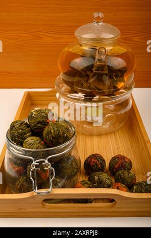Boulettes de thé aux fleurs dans un pot en verre et thé aux fleurs brassées dans une théière en verre sur un plateau en bois sur fond blanc. Gros plan Banque D'Images