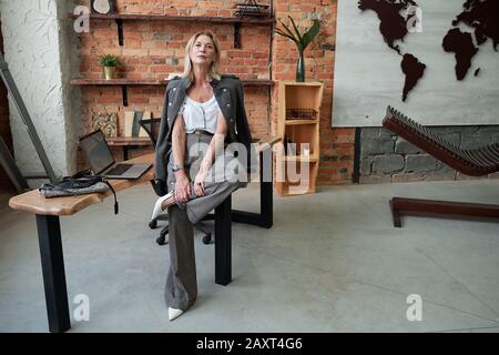 Femme moderne et sûre d'elle avec une tenue élégante assise sur la table et posant pour appareil photo dans le bureau de loft Banque D'Images