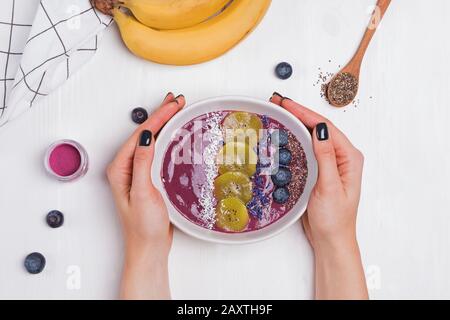 Les mains de la femme tenant le bol à smoothie aux baies d'acai garni de graines, de fleurs et de mûres Banque D'Images