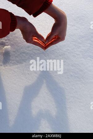 Les mains d'une femme âgée dans une veste rouge jettent une ombre en forme de coeur sur la neige; la lumière douce du soleil coucher du soleil a éclairé une flamme vive entre les doigts Banque D'Images