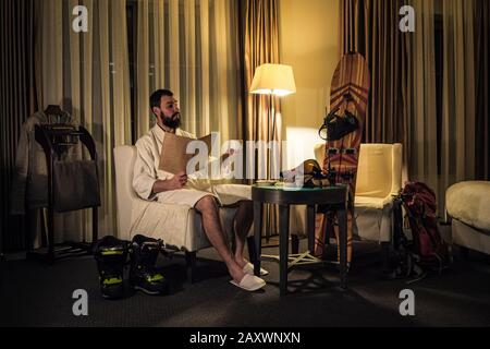 Un snowboarder masculin a un repos dans la chambre d'hôtel en soirée. Un gars dans un peignoir et des pantoufles se trouve dans un plateau de courtoisie, boit du thé et regarde le menu Banque D'Images