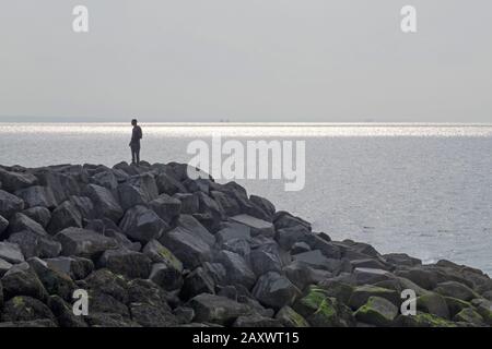 L'homme se tient sur le brise-lames de roche qui donne sur l'océan. Début De Soirée, Été À La Belle Plage, West Sands Selsey, West Sussex, Angleterre. Banque D'Images