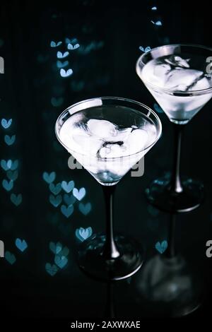 Deux verres à cocktail avec boisson martini avec glace sur fond noir tendance avec lumières bokeh en forme de coeur. Concept de date du soir ou de la nuit. Banque D'Images