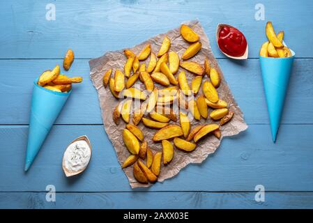 Pommes de terre fraîchement rôties sur papier four et en cônes de papier, sur une table, avec ketchup et crème sure. Vue sur les pommes de terre rustiques sur la table en bois. Banque D'Images