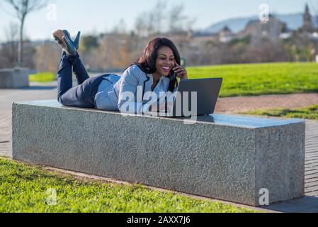 Une femme afro-américaine posée sur un banc à l'aide d'un ordinateur portable et parlant sur un téléphone portable dans un parc extérieur Banque D'Images