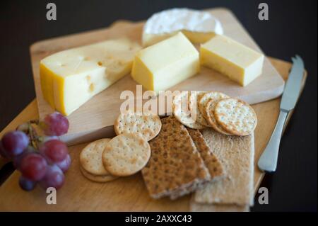 Plateau de fromages et de biscuits avec portions de fromages, biscuits et biscuits à l'eau assortis et un petit bouquet de raisins frais sur fond sombre Banque D'Images