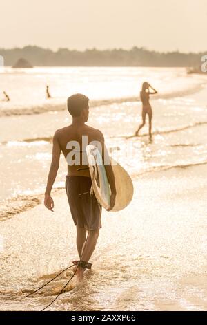 Homme marchant sur la plage tropicale de Midigama, Sri Lankha au soleil avec planche de surf dans ses mains. Banque D'Images