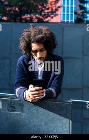 Portrait jeune homme africain à l'aide de son smartphone avec le sourire tout en se penchant sur une clôture à l'extérieur en journée ensoleillée Banque D'Images