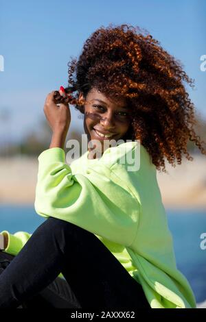 Vue latérale de la belle femme afro curieuse assise sur les rochers de brise-lames riant tout en regardant caméra à l'extérieur Banque D'Images