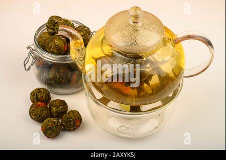 Thé fleuri préparé dans une théière en verre, des boules de thé aux fleurs et un pot de thé sur fond blanc. Gros plan Banque D'Images