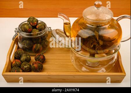 Thé fleuri préparé dans une théière en verre, et un pot avec des boules de thé en fleur sur un plateau en bois sur fond blanc. Gros plan Banque D'Images