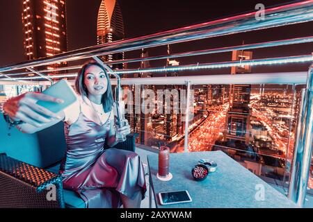 Une fille blogger dans un bar sur le toit prend une photo selfie contre le fond d'une ville de nuit avec des gratte-ciel Banque D'Images