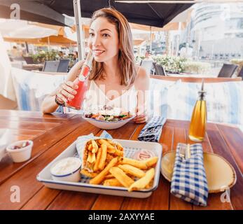 Une jeune fille touristique gaie essaie de déguster une cuisine grecque dans un restaurant local. Sur la table salade traditionnelle Horiatiki et Gyros à Pita Banque D'Images