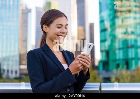 SMS femme utilisant l'application sur smartphone dans le quartier des affaires de la ville. Jeune femme d'affaires utilisant un smartphone souriant portant une veste de costume. Milieu urbain au centre-ville de Vancouver, Canada. Banque D'Images