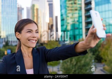 Femme d'affaires prenant une photo selfie à l'aide de l'application smartphone sur smartphone pour les réseaux sociaux. Jeune femme d'affaires en utilisant un smartphone souriant heureux. Vancouver, Canada à l'extérieur. Femme professionnelle urbaine. Banque D'Images