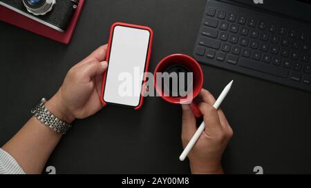 Prise de vue aérienne d'un photographe masculin tenant un smartphone maquette tout en travaillant sur un lieu de travail élégant avec un stylet, une tablette et un appareil photo sur un bureau noir Banque D'Images