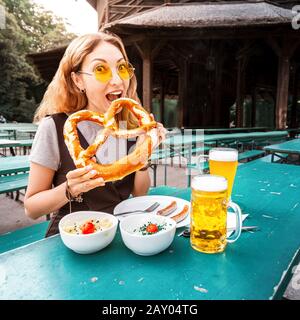 Une fille asiatique mangeant du Pretzel traditionnel et buvant de la bière bavaroise fraîche dans le jardin à bière de Munich. Le concept de festival alimentaire traditionnel et de tourisme Banque D'Images