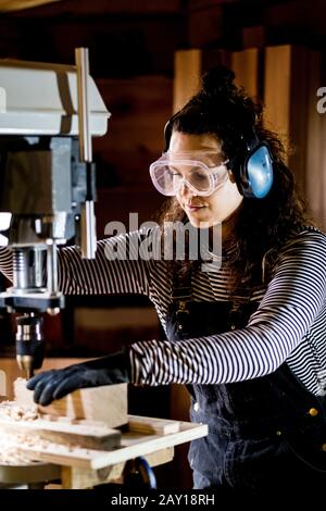 Femme avec cheveux longs marron portant des salopettes, des lunettes de sécurité et des protections d'oreilles debout dans l'atelier de bois, à l'aide d'une perceuse électrique. Banque D'Images