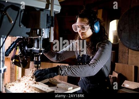 Femme avec cheveux longs marron portant des salopettes, des lunettes de sécurité et des protections d'oreilles debout dans l'atelier de bois, à l'aide d'une perceuse électrique. Banque D'Images