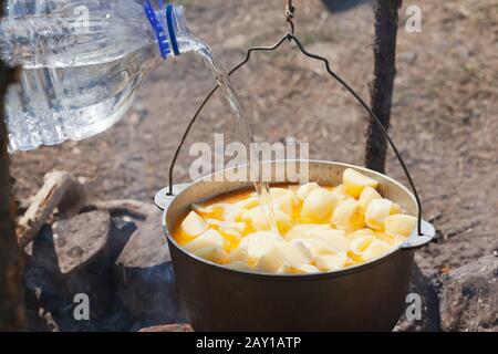 Ajouter de l'eau de grande bouteille en plastique dans un chaudron noir avec pomme de terre sur feu ouvert, repas de camping Banque D'Images