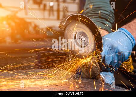 Les côtés rapprochés volent les étincelles lumineuses de la machine à meuleuse angulaire. Un jeune soudeur mâle dans des gants de travail bleus grince un produit métallique avec angle Banque D'Images