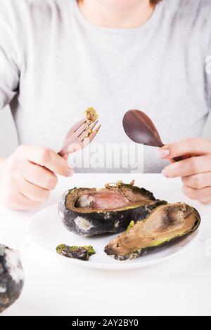 Un homme sans visage est assis à une table, avec un couteau et une fourche dans ses mains devant une plaque d'avocat pourri. Banque D'Images