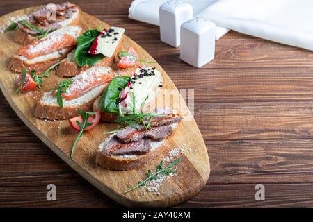 Sandwichs au poisson rouge, au bœuf en suvit et au fromage doux. Garnies de tranches de microvert, d'arugula et de tomates. Sur un plateau ovale en bois espace de copie Banque D'Images