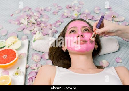 Femme avec masque facial en argile rose dans un spa de beauté. En arrière-plan pétales de fleurs et fruits frais, tranches de pamplemousse, pomme, citron. Banque D'Images