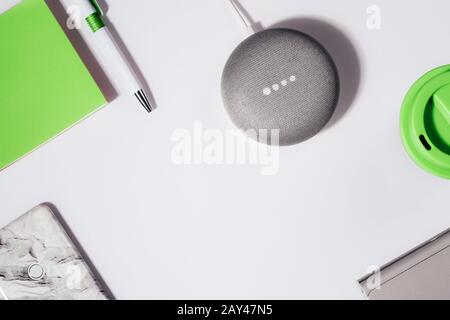 Concept d'assistant numérique. Enceinte plate avec éclairage LED activée avec smartphone court, bloc-notes vert, stylo et tasse à café réutilisable. Banque D'Images