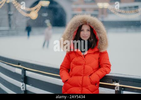 Jeune femme séduisante dans une veste chaude rouge, se tient près de la patinoire, allant skate, garde les mains dans les poches, regarde avec un sourire doux directement à l'appareil photo, s Banque D'Images