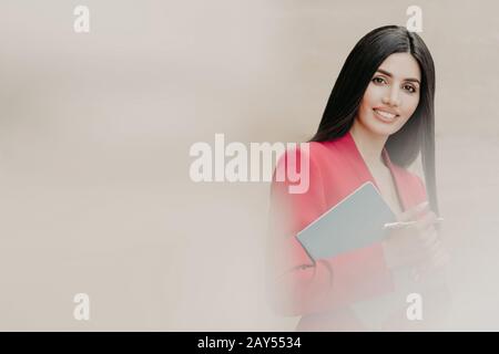 Photo horizontale d'une jolie femme brunette avec une bonne expression faciale, vêtue de vêtements formels, tient le carnet et le stylo, a des cheveux droits noirs, sta Banque D'Images