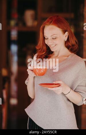 Jolie jeune femme européenne avec des cheveux rouges et frisés, vêtus d'une tenue décontractée, boit du café ou du thé, a du temps libre, sourit doucement, a de la forme, bein Banque D'Images