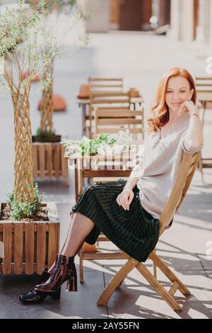 Photo verticale d'une jolie femme aux cheveux rouges assise sur la chaise, attend pour commander dans un café extérieur, porte un pull, une jupe et des chaussures, regarde l'appareil photo avec satisfaction Banque D'Images