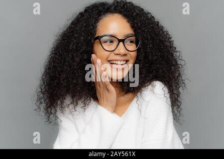 La tête de la belle peau sombre modèle femme touche doucement joue, a le sourire charmant sur le visage, semble heureux loin, a curieusement Afro cheveux, porte spect Banque D'Images