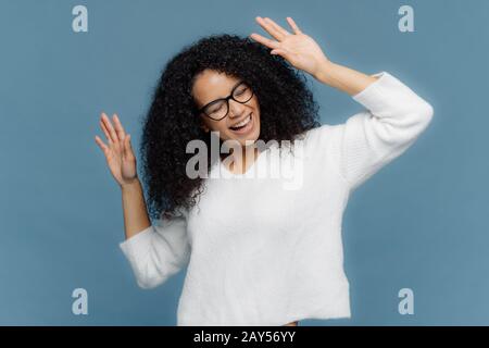 Concept de personnes et de joie. Optimiste enchantée Afro American femme lève les mains, danse à la musique forte, être surjoyé, porte un pull blanc décontracté, isola Banque D'Images