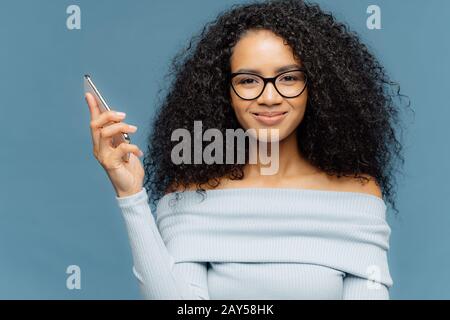 Photo d'une femme agréable avec des cheveux raillés, sourires heureux, tient un téléphone portable moderne pour discuter en ligne, porte un pull à la mode, semble traigh Banque D'Images