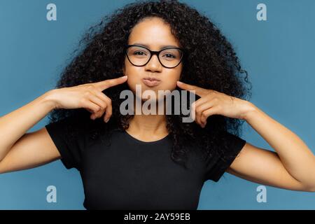 Drôle de femme à peau foncée avec des cheveux frisés, appuie sur les joues, retient la respiration, souffle les joues, porte des lunettes optiques et un t-shirt décontracté, isolé sur bleu bac Banque D'Images