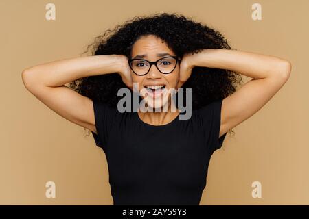 Jeune femme aux cheveux bouclés Afro couvre les oreilles, les becs en tant que demande être calme, ne pas se tenir au bruit, garde la bouche ouverte, porte des lunettes et t sh noir décontracté Banque D'Images