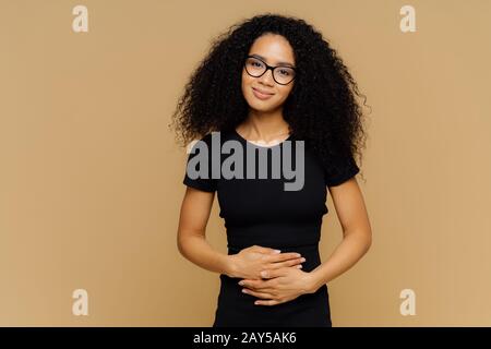 Heureuse Afro American femme avec des cheveux rabouclés, garde les mains sur le ventre, se sent bien après avoir mangé un délicieux dîner nutritif, regarde heureux à l'appareil photo. Jeune Banque D'Images