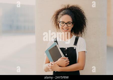 Adorable fille aux cheveux bouclés porte un t-shirt blanc décontracté et des combinaisons, tient un bloc-notes ou un manuel, regarde par des lunettes transparentes, des stands Banque D'Images