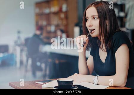 Pensive femme blogger écrit dans des idées de bloc-notes pour la publication dans le blog, assis contre l'intérieur de café, garde le stylo à la bouche, boissons café concentré loin, dr Banque D'Images