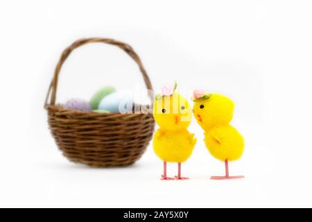 Deux jouets jaunes de pâques aux chapeaux de roses chuchotant devant un panier flou sur fond blanc. Banque D'Images