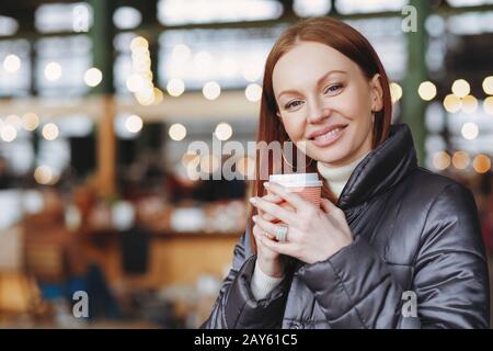 Photo horizontale d'une jolie femme au sourire toothy, conserve une tasse de boisson en papier, aime le café aromatique, porte une veste à la mode, pose sur le dos flou Banque D'Images