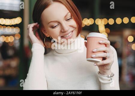 La photo courte d'une femme agréable pose dans un café extérieur, contient une tasse de café en papier, vêtue d'une tenue blanche, sourit avec plaisir, a parlé avec ses collègues Banque D'Images