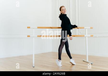 La jeune femme aux cheveux rouges mince se tient sur les orteils de la pointe, étant en bonne forme de corps, s'entraîne tous les jours, porte un sweat-shirt noir, des leggings et wh Banque D'Images