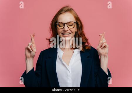 La photo d'une femme d'affaires heureuse croise les doigts pour la bonne chance, croit que tout va bien, prie sur fond rose, a le sourire toothy, porte des spécifications Banque D'Images