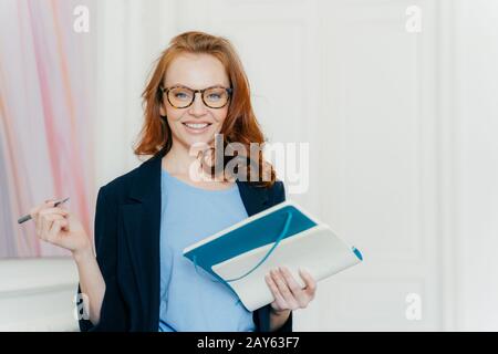 Satisfait freelancer écrit le futur plan pour la semaine prochaine, prépare la recherche pour le démarrage, fait des notes dans le bloc-notes, tient le stylo, regarde avec joie à l'appareil photo, dr Banque D'Images