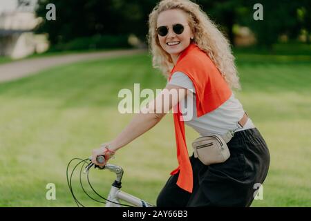 La photo horizontale d'une heureuse femme qui fait du vélo, garde les mains sur le guidon, semble active et rafraîchie, vêtue de façon décontractée, porte des nuances, pose contre Banque D'Images