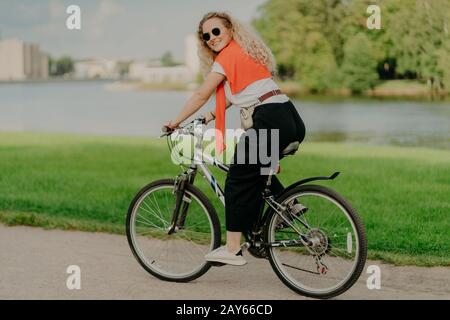 Heureuse jeune femme bénéficie d'un nouvel itinéraire à vélo, des promenades au bord du lac, de la pelouse verte et des bâtiments loin de distance, porte des teintes d'été, tenue décontractée Banque D'Images